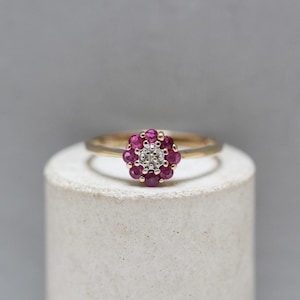 Puede incluir: Anillo de oro con un diseño floral, engastado con piedras preciosas de color rojo intenso y un grupo central de piedras claras. El anillo se exhibe sobre un soporte cilíndrico blanco.