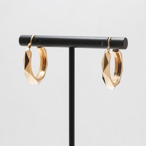 Op de afbeelding: Een paar gouden creolen met een geometrisch ontwerp. De oorbellen worden gepresenteerd op een zwarte standaard.