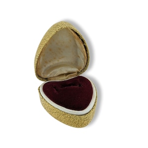 Puede incluir: Una caja de anillo en forma de corazón con un exterior de color dorado y un interior de terciopelo rojo. La caja está abierta y el forro de terciopelo es visible.