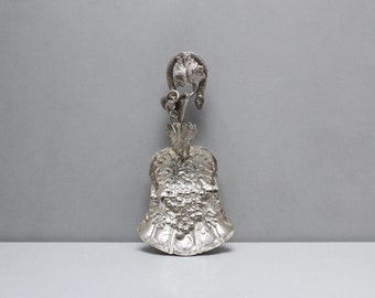 May include: A silver sugar scoop with an ornate design featuring a grapevine and a cluster of grapes. The handle is shaped like a vine with leaves.