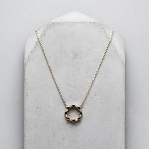 Può includere: Una collana dorata con un pendente circolare ornato da piccole pietre preziose rosso scuro. La collana è esposta su una superficie grigia strutturata, evidenziando il suo design delicato. La catena è sottile ed elegante.