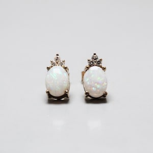 May include: A pair of stud earrings featuring oval-shaped white opals with iridescent flashes of color. Each opal is set in a gold-toned metal prong setting, topped with a small cluster of three clear, sparkling round-cut accent stones.