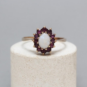 May include: A gold ring featuring a central oval opal gemstone surrounded by a halo of small, dark red gemstones. The ring is set on a light-colored, cylindrical surface against a soft gray background.