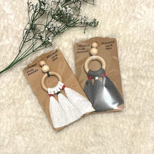 May include: Two macrame essential oil diffusers. One has white tassels with red accents, the other has grey tassels. Each has a wooden ring and beads. The packaging reads "Diffuseur d'huile essentielle" and "Macramé 100% coton."