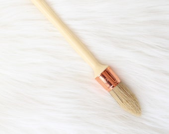 May include: A wooden-handled paint brush with a copper ferrule and natural bristles. The brush is on a white fluffy surface.