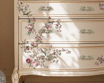 May include: A cream-coloured three-drawer chest featuring gold trim and a floral decal with pink roses and small birds. The chest has curved legs and ornate metal drawer pulls. A white vase filled with white flowers sits on top of the chest.