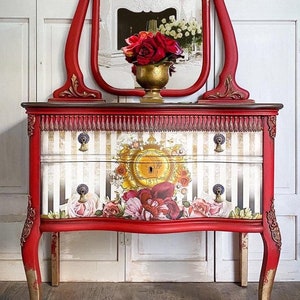 May include: A red vanity table with a gold-trimmed mirror and floral patterned drawers. The table has a distressed finish and features ornate details. The mirror is oval-shaped and has a beveled edge. The drawers are decorated with a floral pattern and have gold knobs. The table is sitting on a wooden floor.