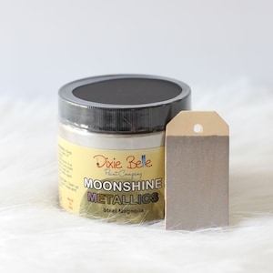 K&ouml;nnte beinhalten: Ein runder Farbtopf der Dixie Belle Paint Company Moonshine Metallics in der Farbe Steel Magnolia mit schwarzem Deckel. Neben dem Topf liegt ein rechteckiges Holzschild, das mit einer Probe der metallisch grauen Farbe bemalt ist und ein Loch hat.