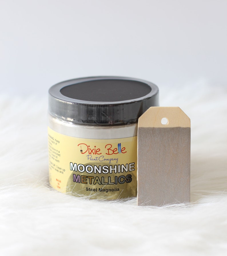 K&ouml;nnte beinhalten: Ein runder Farbtopf der Dixie Belle Paint Company Moonshine Metallics in der Farbe Steel Magnolia mit schwarzem Deckel. Neben dem Topf liegt ein rechteckiges Holzschild, das mit einer Probe der metallisch grauen Farbe bemalt ist und ein Loch hat.