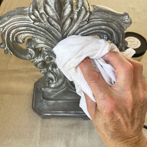 May include: A detailed shot of a silver-colored decorative lamp base with ornate floral designs. A white cloth is used to clean the lamp base. The lamp base is on a beige surface.