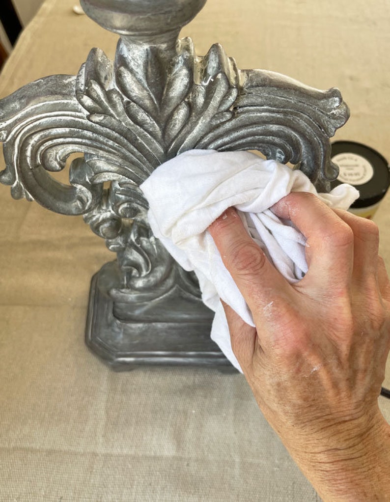May include: A detailed shot of a silver-colored decorative lamp base with ornate floral designs. A white cloth is used to clean the lamp base. The lamp base is on a beige surface.