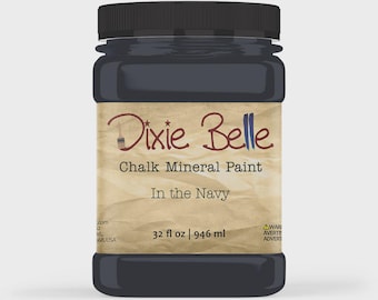 May include: A dark navy blue chalk mineral paint in a plastic jar with a black screw-top lid. The label features the text Dixie Belle Chalk Mineral Paint In the Navy and indicates a volume of 946 ml or 32 fl oz on a tan, textured background.