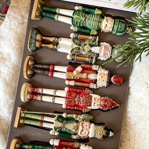 May include: A silicone mold for making nutcracker figurines. The mold is gray and features eight different nutcracker designs in various colors, including red, green, gold, and white. The mold is labeled "Decor Moulds Nutcracker Magic" and "Item No: 670382 Nutcracker Magic".
