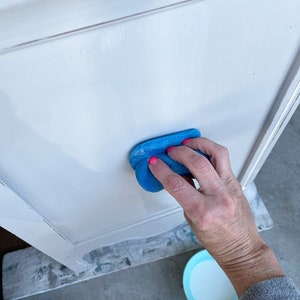Puede incluir: Una mano frota una esponja azul sobre una superficie pintada de blanco. La esponja se utiliza para alisar la pintura y crear un acabado uniforme.