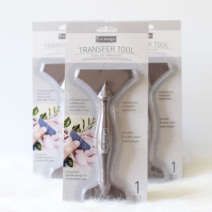 May include: A gray transfer tool with a handle and a flat, rectangular surface. The tool is packaged in a clear plastic sleeve with the text "re-design Transfer Tool" and "Outil de Transfert Herramienta de Transferencia".