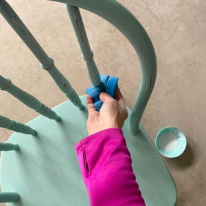 Puede incluir: Una mano se muestra usando una esponja de lijar azul para desgastar una silla pintada de azul claro. La silla tiene un respaldo redondeado y un asiento de madera.