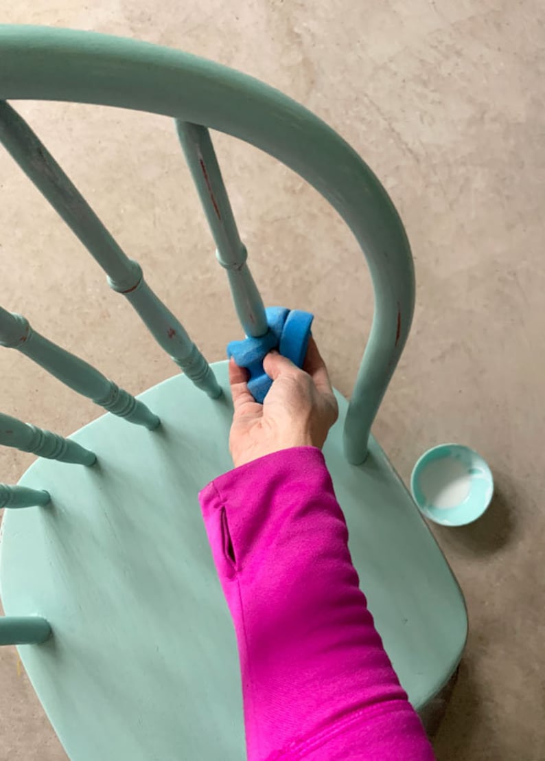 Puede incluir: Una mano se muestra usando una esponja de lijar azul para desgastar una silla pintada de azul claro. La silla tiene un respaldo redondeado y un asiento de madera.