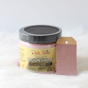 K&ouml;nnte beinhalten: Ein Farbtopf von Dixie Belle Paint Company Moonshine Metallics in der Farbe Rozay mit schwarzem Deckel und gelbem Etikett mit schwarzer Schrift. Neben dem Topf liegt ein rechteckiges Holzschild, das mit der gleichen metallisch rosa Farbe bemalt ist.