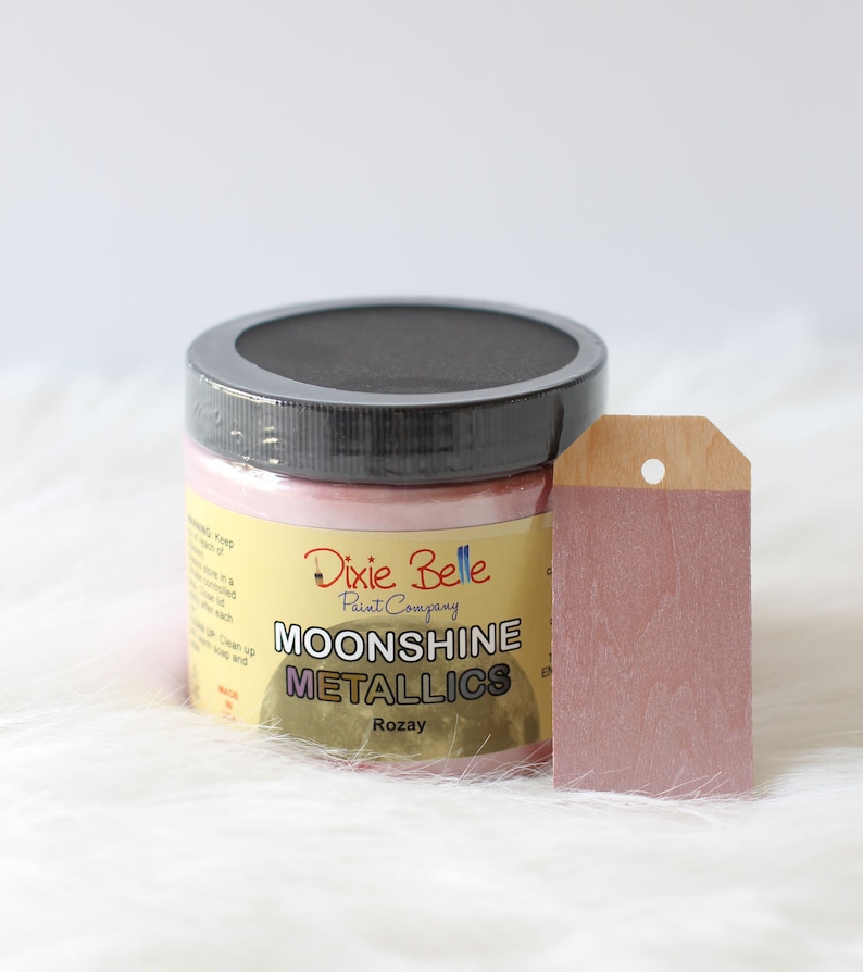 K&ouml;nnte beinhalten: Ein Farbtopf von Dixie Belle Paint Company Moonshine Metallics in der Farbe Rozay mit schwarzem Deckel und gelbem Etikett mit schwarzer Schrift. Neben dem Topf liegt ein rechteckiges Holzschild, das mit der gleichen metallisch rosa Farbe bemalt ist.