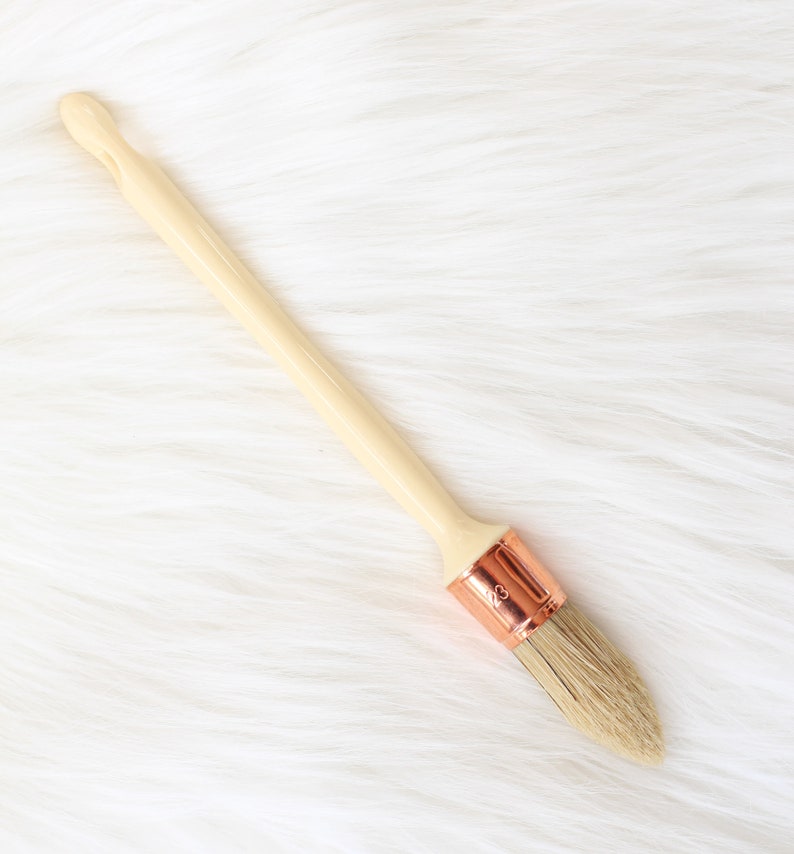 May include: A wooden-handled paint brush with a copper ferrule and natural bristles. The brush is on a white fluffy surface.