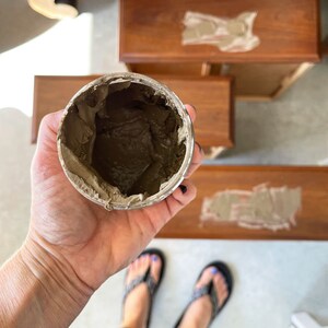 Pu&ograve; includere: Un primo piano di un piccolo barattolo aperto di stucco per legno marrone. Il barattolo &egrave; tenuto da una mano. Lo stucco per legno viene utilizzato per riempire fessure e buchi nel legno.