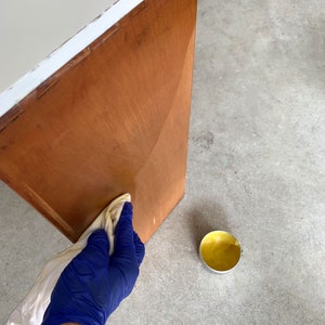 Puede incluir: Una persona con un guante azul aplica cera para madera a una puerta de armario de madera con un borde blanco. La puerta del armario es de color marr&oacute;n c&aacute;lido. Un peque&ntilde;o bote abierto de cera amarilla est&aacute; cerca, sobre una superficie de hormig&oacute;n.