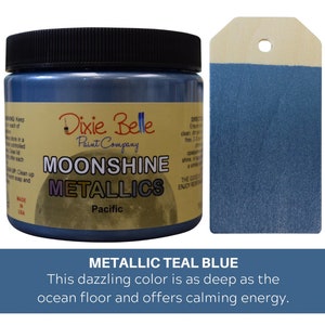 K&ouml;nnte beinhalten: Ein Farbtopf von Dixie Belle Moonshine Metallics in der Farbe Pacific mit schwarzem Deckel und einem Etikett mit der Aufschrift Dixie Belle Paint Company Moonshine Metallics Pacific. Daneben liegt ein h&ouml;lzernes Etikett, das in metallischem Blaugr&uuml;n gestrichen ist.