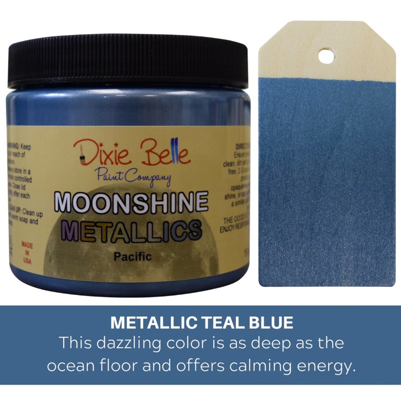 K&ouml;nnte beinhalten: Ein Farbtopf von Dixie Belle Moonshine Metallics in der Farbe Pacific mit schwarzem Deckel und einem Etikett mit der Aufschrift Dixie Belle Paint Company Moonshine Metallics Pacific. Daneben liegt ein h&ouml;lzernes Etikett, das in metallischem Blaugr&uuml;n gestrichen ist.