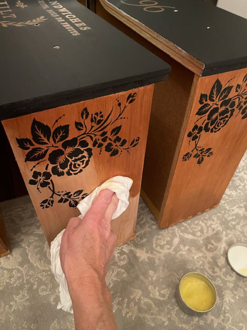 Puede incluir: Dos mesas auxiliares de madera con tapas negras y plantillas florales. Las mesas son de un c&aacute;lido tono de madera con dise&ntilde;os florales negros. Una mesa tiene texto en la parte superior. Una mano limpia el lateral de una mesa con un pa&ntilde;o.