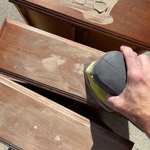 Pu&ograve; includere: Primo piano di una mano che usa una levigatrice elettrica per lisciare la superficie di un cassetto in legno. Il cassetto &egrave; in legno scuro e ha una finitura marrone chiaro. La levigatrice &egrave; gialla e nera e ha un'impugnatura in gomma nera.