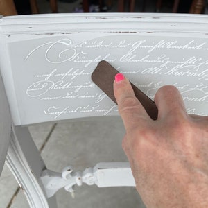 May include: A hand rubs a brown tool over a white painted surface with white script lettering that reads "Dies as unter der Gerichtsbarkeit den Oberlandese nach erfolgtert und Kittengut von den vier G Consens und nach - "