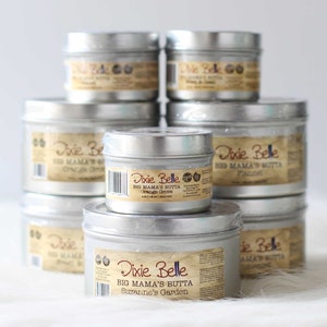May include: A stack of silver metal tins of Dixie Belle Big Mama's Butta. Each tin has a brown label with product information. The labels include the product names: Orange Grove, Flannel, and Suzanne's Garden. The tins are arranged on a white surface.