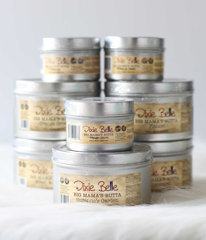 Puede incluir: Una pila de latas de metal plateado de Dixie Belle Big Mama's Butta. Cada lata tiene una etiqueta marr&oacute;n con informaci&oacute;n del producto. Las etiquetas incluyen los nombres de los productos: Orange Grove, Flannel y Suzanne's Garden. Las latas est&aacute;n dispuestas sobre una superficie blanca.