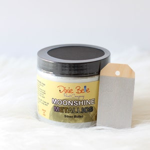 K&ouml;nnte beinhalten: Ein runder Kunststofftiegel mit Moonshine Metallics Farbe der Dixie Belle Paint Company in der Farbe Silver Bullet. Der Tiegel hat einen schwarzen Deckel und ein gelbes Etikett mit schwarzer Schrift. Daneben liegt ein silberfarbenes Holzschild.