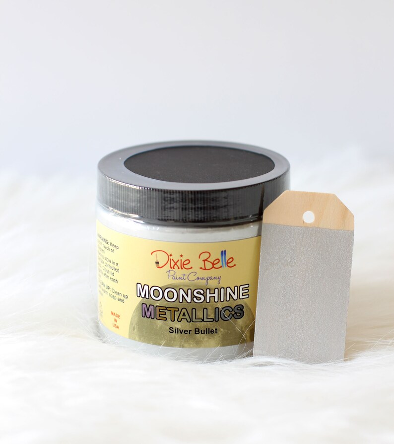 K&ouml;nnte beinhalten: Ein runder Kunststofftiegel mit Moonshine Metallics Farbe der Dixie Belle Paint Company in der Farbe Silver Bullet. Der Tiegel hat einen schwarzen Deckel und ein gelbes Etikett mit schwarzer Schrift. Daneben liegt ein silberfarbenes Holzschild.