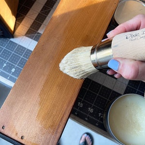 Puede incluir: Una superficie de madera est&aacute; siendo tratada con un cepillo y cera. El cepillo, etiquetado como "Dixie Belle Paint Company", aplica cera a la madera marr&oacute;n claro. Una peque&ntilde;a lata de cera est&aacute; cerca. La madera tiene un borde oscuro y est&aacute; sobre una esterilla de corte.