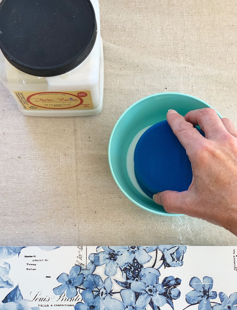 Puede incluir: Un tarro de pl&aacute;stico blanco con una tapa negra etiquetado como "Dixie Belle 2 oz Clear Coat Satin" y una esponja azul en un taz&oacute;n azul claro.