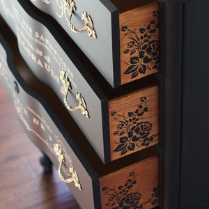 Puede incluir: C&oacute;moda pintada de negro con tres cajones abiertos. Los cajones tienen tiradores dorados y dise&ntilde;os florales en el interior. La parte delantera de los cajones tiene letras doradas. La c&oacute;moda est&aacute; sobre un suelo de madera.