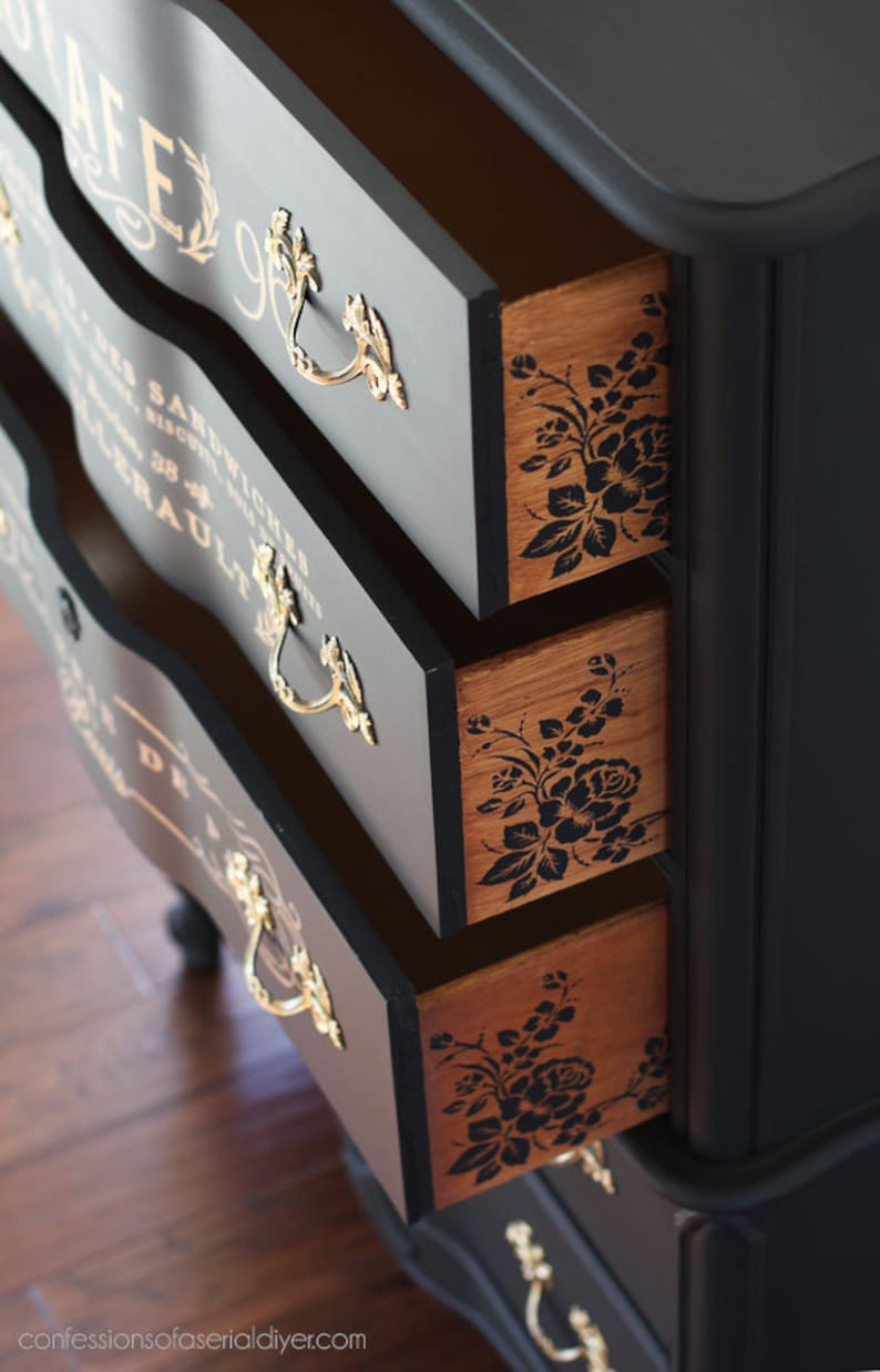 Puede incluir: C&oacute;moda pintada de negro con tres cajones abiertos. Los cajones tienen tiradores dorados y dise&ntilde;os florales en el interior. La parte delantera de los cajones tiene letras doradas. La c&oacute;moda est&aacute; sobre un suelo de madera.