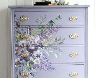 Puede incluir: Una cómoda lavanda con cinco cajones y tiradores de cajones ovalados dorados. La cómoda está decorada con una calcomanía floral con flores moradas, verdes y blancas.
