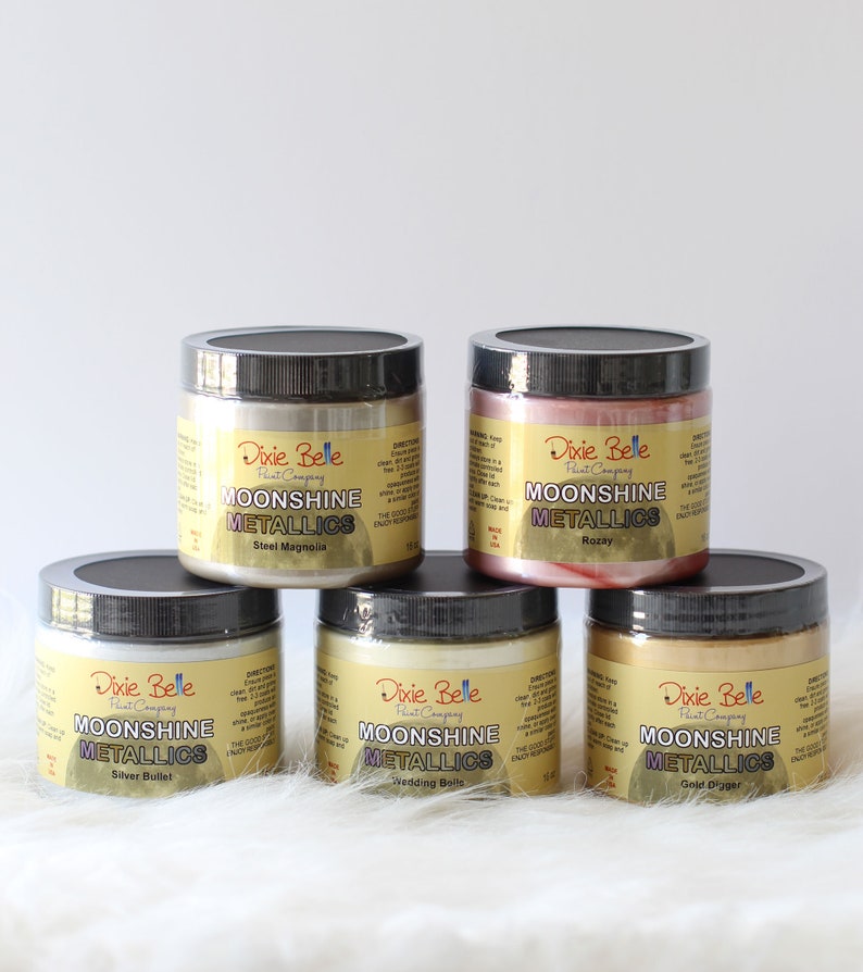K&ouml;nnte beinhalten: F&uuml;nf Farbdosen von Dixie Belle Moonshine Metallics mit schwarzen Deckeln. Die Etiketten tragen den Text Moonshine Metallics und die Farbnamen: Steel Magnolia, Rozay, Silver Bullet, Wedding Belle und Gold Digger. Jede Dose enth&auml;lt 473 ml.