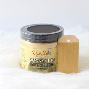 K&ouml;nnte beinhalten: Ein runder Farbtopf von Dixie Belle Paint Company Moonshine Metallics Gold Digger mit einem schwarzen Deckel. Das Etikett zeigt den Markennamen und den Produktnamen in schwarzer und wei&szlig;er Schrift. Ein mit der Goldfarbe bemalter Holzanh&auml;nger steht daneben.