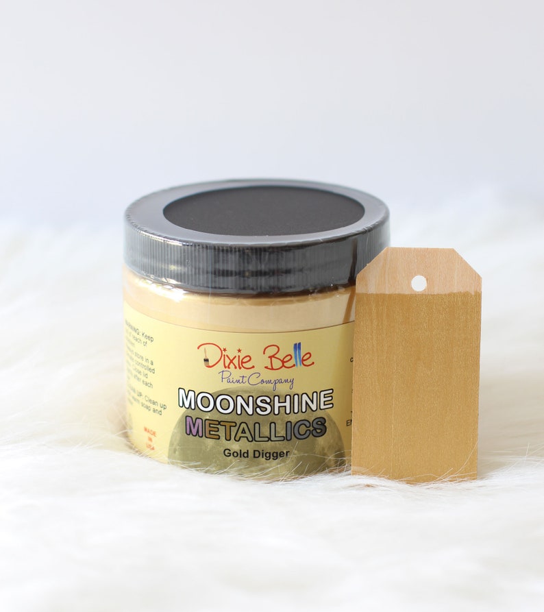 K&ouml;nnte beinhalten: Ein runder Farbtopf von Dixie Belle Paint Company Moonshine Metallics Gold Digger mit einem schwarzen Deckel. Das Etikett zeigt den Markennamen und den Produktnamen in schwarzer und wei&szlig;er Schrift. Ein mit der Goldfarbe bemalter Holzanh&auml;nger steht daneben.
