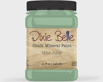 May include: A plastic jar of Dixie Belle Chalk Mineral Paint in the colour Mint Julep. The label features a tan, textured background with the brand name, product type, and the volume 946 ml. The jar has a black screw-top lid and contains mint green paint.