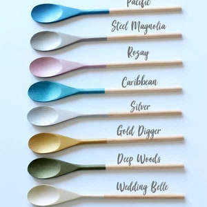 K&ouml;nnte beinhalten: Acht Holzl&ouml;ffel sind vertikal angeordnet, jeder mit einer lackierten Laffe in Metallic-Farben wie Blau, Silber, Rosa, T&uuml;rkis, Gold, Gr&uuml;n und Wei&szlig;. Die Griffe sind aus naturbelassenem Holz. Die Beschriftungen lauten Pacific, Steel Magnolia, Rozay, Caribbean, Silver, Gold Digger, Deep Woods und Wedding Belle.