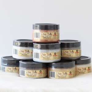 May include: A collection of Dixie Belle glaze jars in various colors, including Copper Bronze, Whitewash, Hi-Ho Silver, and Gold Glimmer. Each jar has a black lid and a label with product information. The jars are stacked on a white surface.