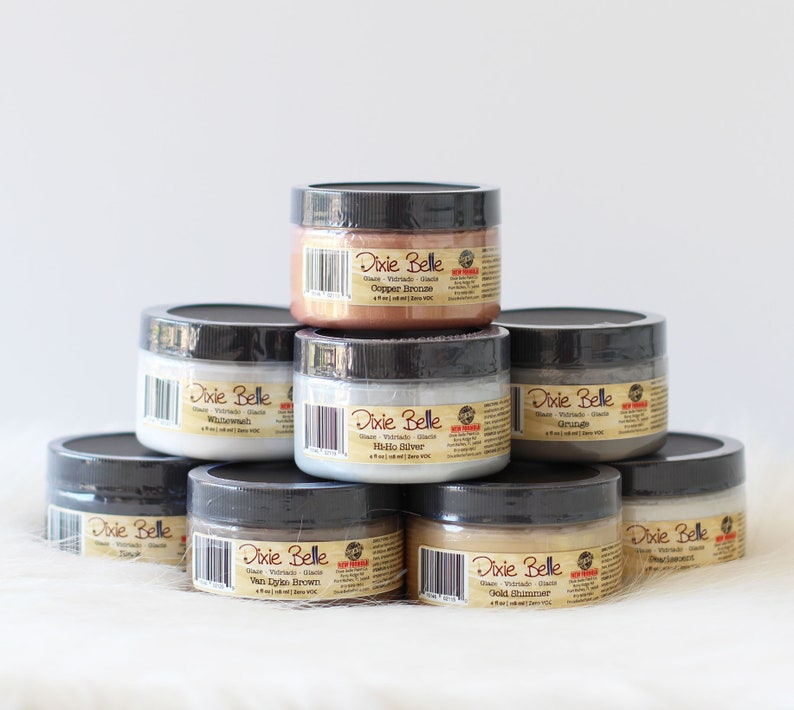 May include: A collection of Dixie Belle glaze jars in various colors, including Copper Bronze, Whitewash, Hi-Ho Silver, and Gold Glimmer. Each jar has a black lid and a label with product information. The jars are stacked on a white surface.