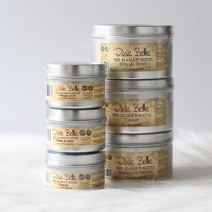 Puede incluir: Una pila de latas de metal plateado de Dixie Belle Big Mama's Butta. Las etiquetas son de color beige con texto negro, que enumeran aromas como Orange Grove, Flannel, Fresh & Clean y Suzanne's Garden. Las latas est&aacute;n sobre una superficie blanca.