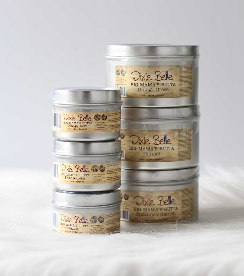Puede incluir: Una pila de latas de metal plateado de Dixie Belle Big Mama's Butta. Las etiquetas son de color beige con texto negro, que enumeran aromas como Orange Grove, Flannel, Fresh & Clean y Suzanne's Garden. Las latas est&aacute;n sobre una superficie blanca.