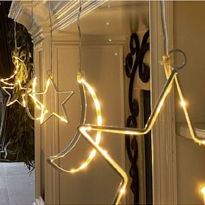 May include: A string of lights shaped like stars and crescent moons. The lights are white and gold and are strung together with clear wire. The lights are hanging from a ceiling.